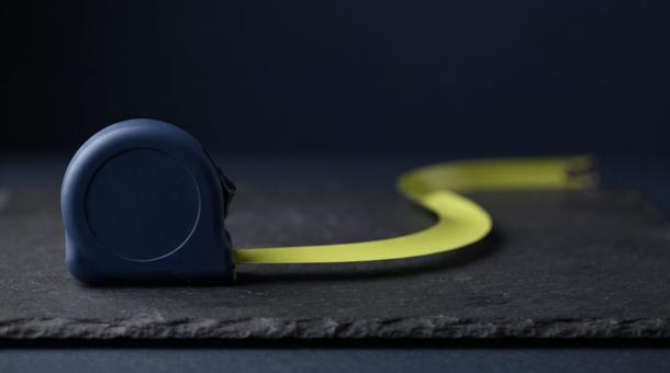 A tape measure with a navy case on dark slate, its chartreuse blade pulled out in a long curling arc.