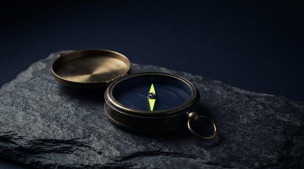 An open pocket compass on dark slate, its case in dark brass and its needle bright chartreuse.