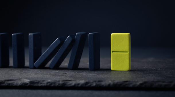 A line of navy dominoes on dark slate, the first few already toppling, the last one chartreuse and still standing.