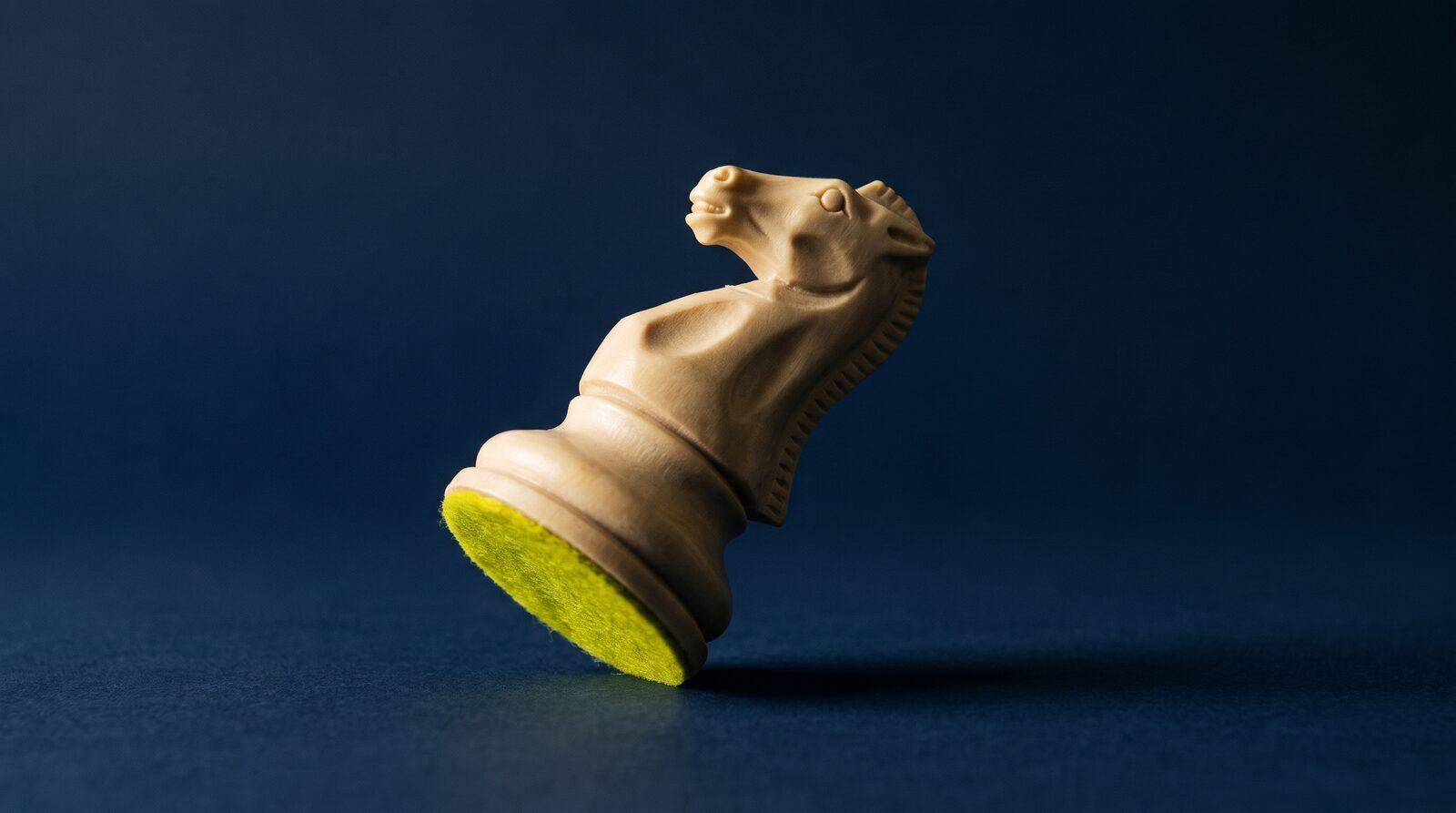 A pale bone chess knight tipped past its balance point on navy stone, caught mid-fall, acid yellow-green felt showing on its lifted base