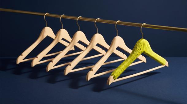 A row of cream wooden coat hangers on a slim brass rail against smooth navy, every hanger empty, the one at the end wound with acid yellow-green thread.