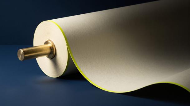 A long blank cream paper roll unspooling from a brass spindle and running away across smooth navy into shadow, one acid yellow-green stripe along its edge.