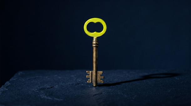 A single door key standing upright on its teeth on navy stone, the bow of the key in acid yellow-green, casting one long shadow.
