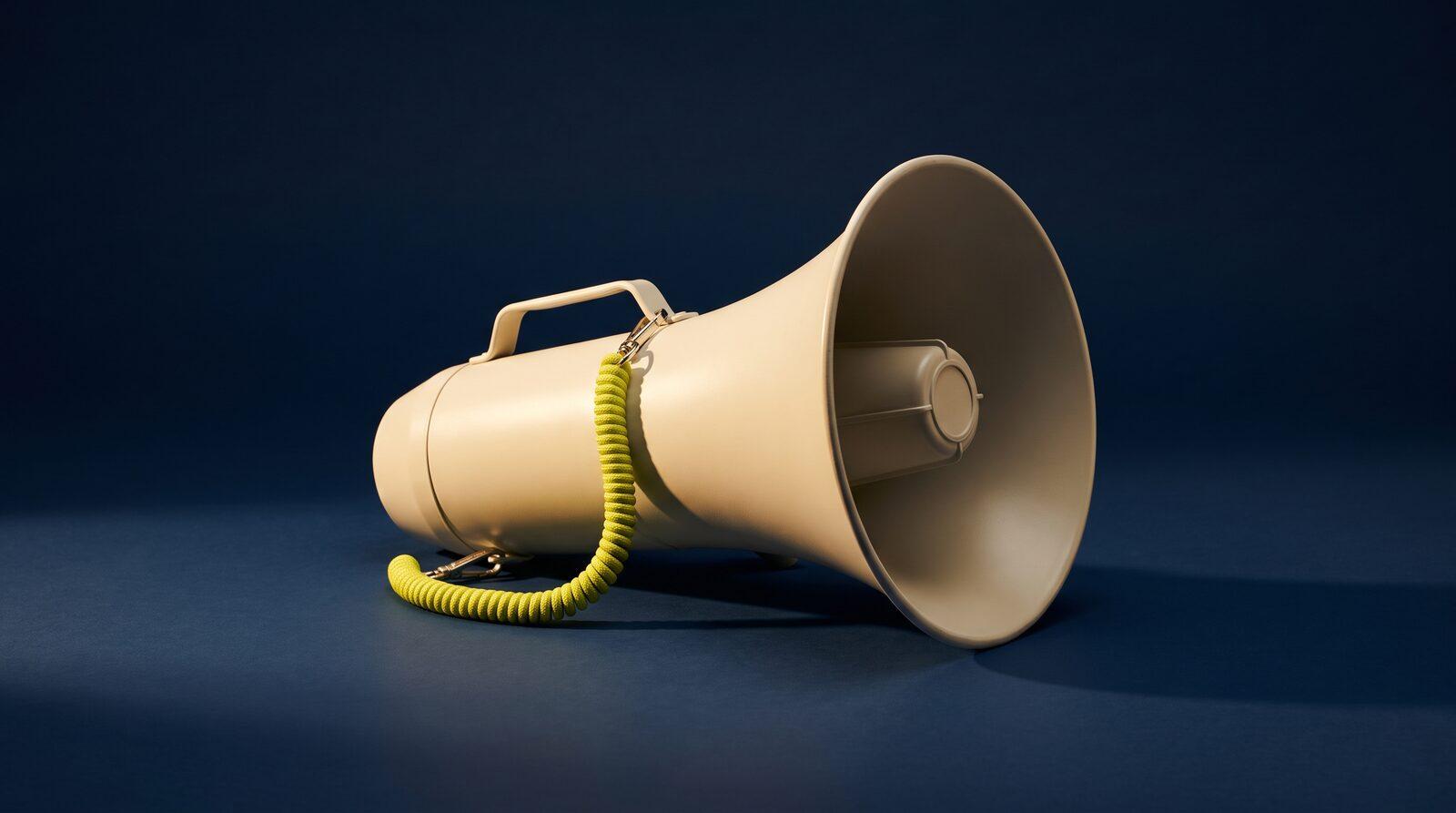 A cream metal megaphone lying on its side on a smooth navy surface, a coiled acid yellow-green cord looped from its handle, lit hard from one side