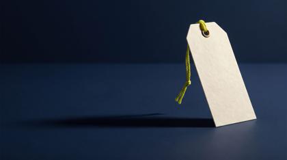 A blank cream price tag propped upright on navy stone, nothing written on it, an acid yellow-green thread knotted through its brass eyelet, casting a long shadow