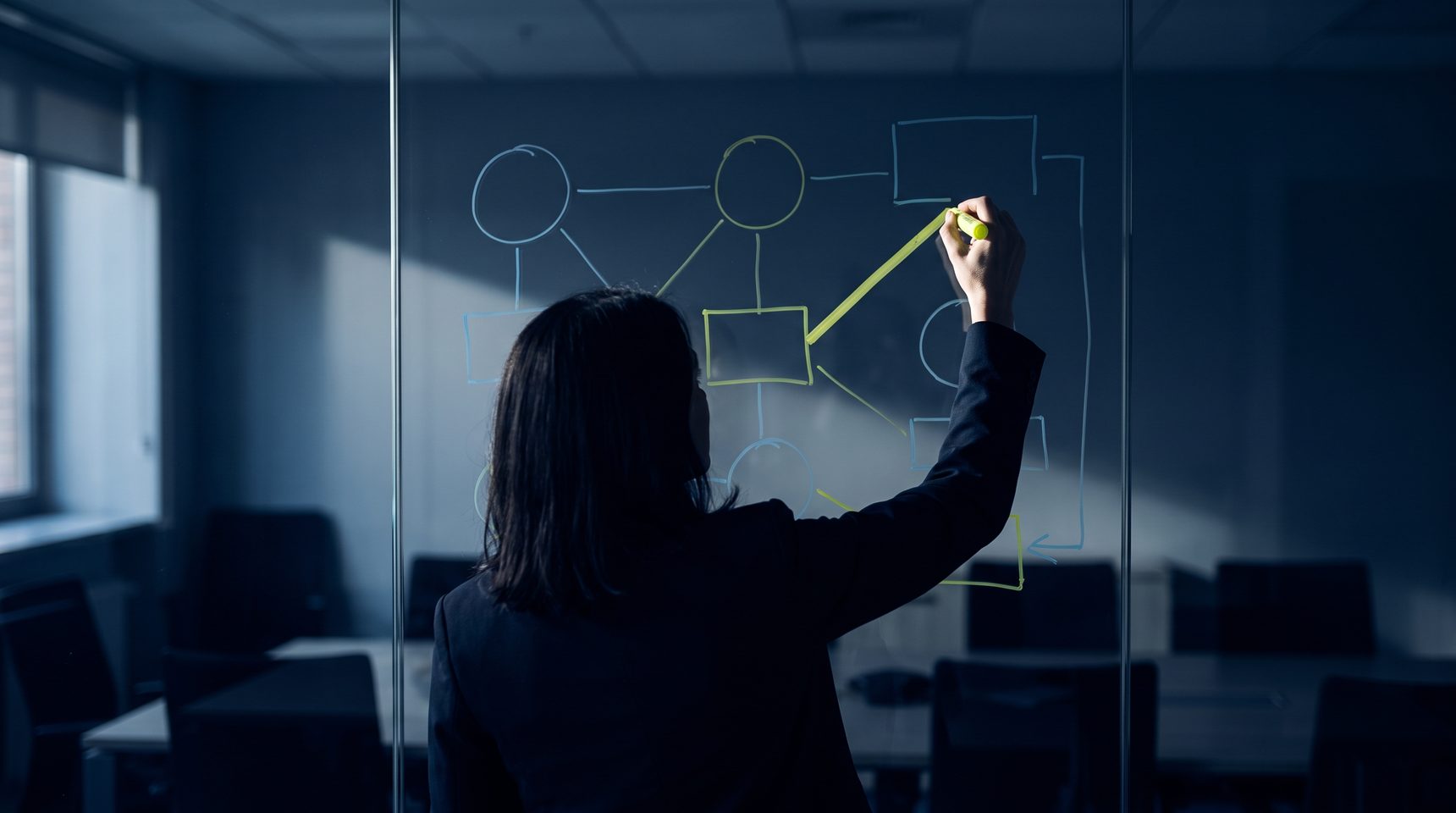 A person seen from behind marks one connecting line in yellow-green on a glass wall covered in hand-drawn boxes and arrows, in a darkened meeting room.