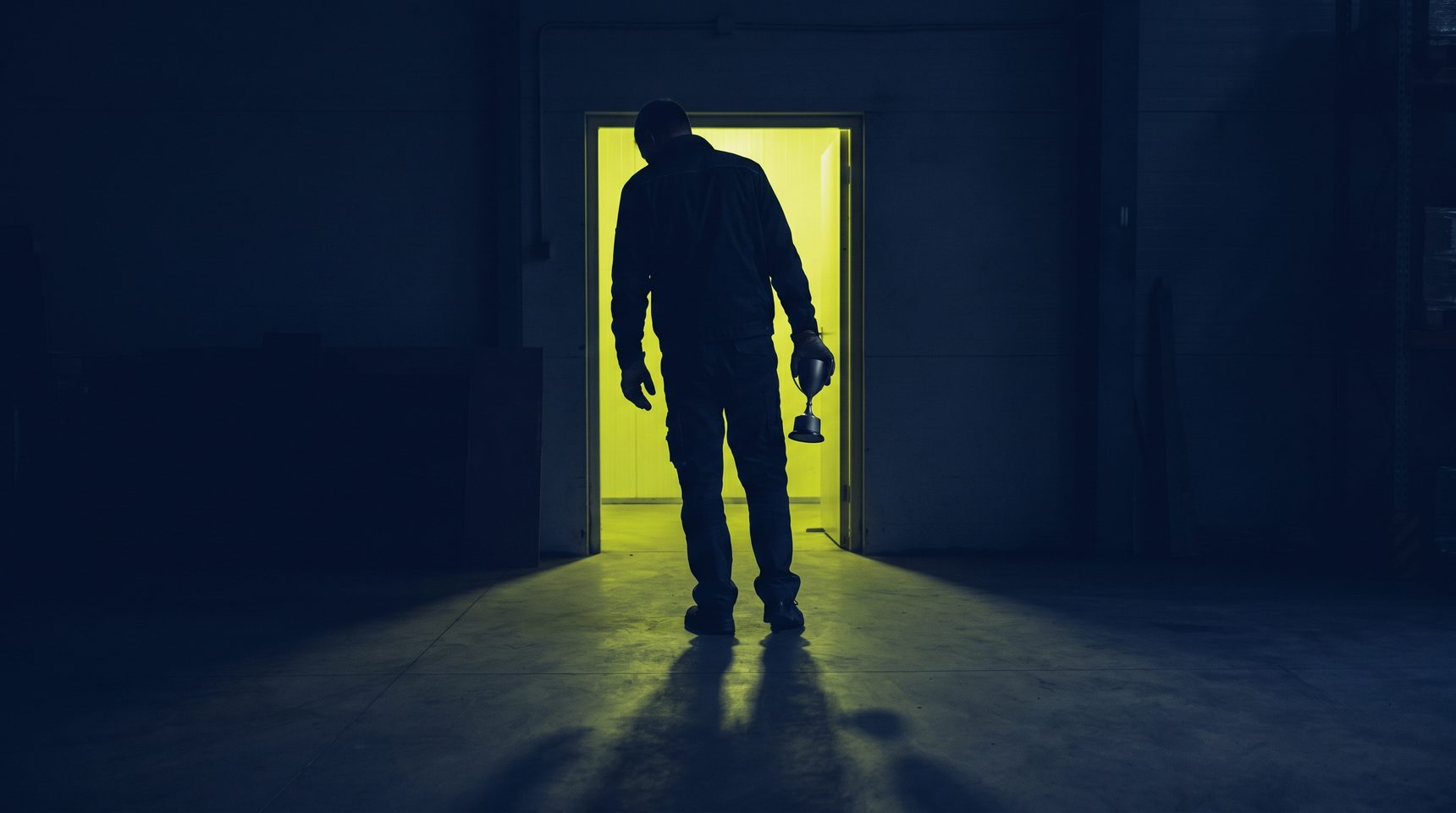 A figure seen from behind in silhouette holds a small plain trophy at their side, standing in a dark room before an open doorway filled with bright yellow-green light.