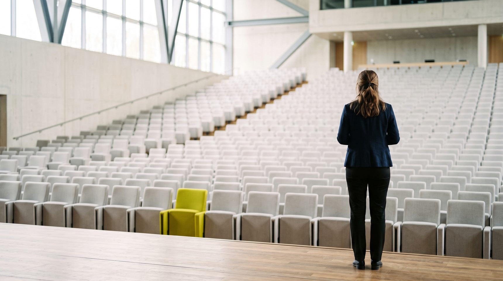 A woman stands with her back to the camera at the edge of a bright auditorium stage before the audience arrives, rows of empty pale seats rising in front of her and a single yellow-green seat among them.