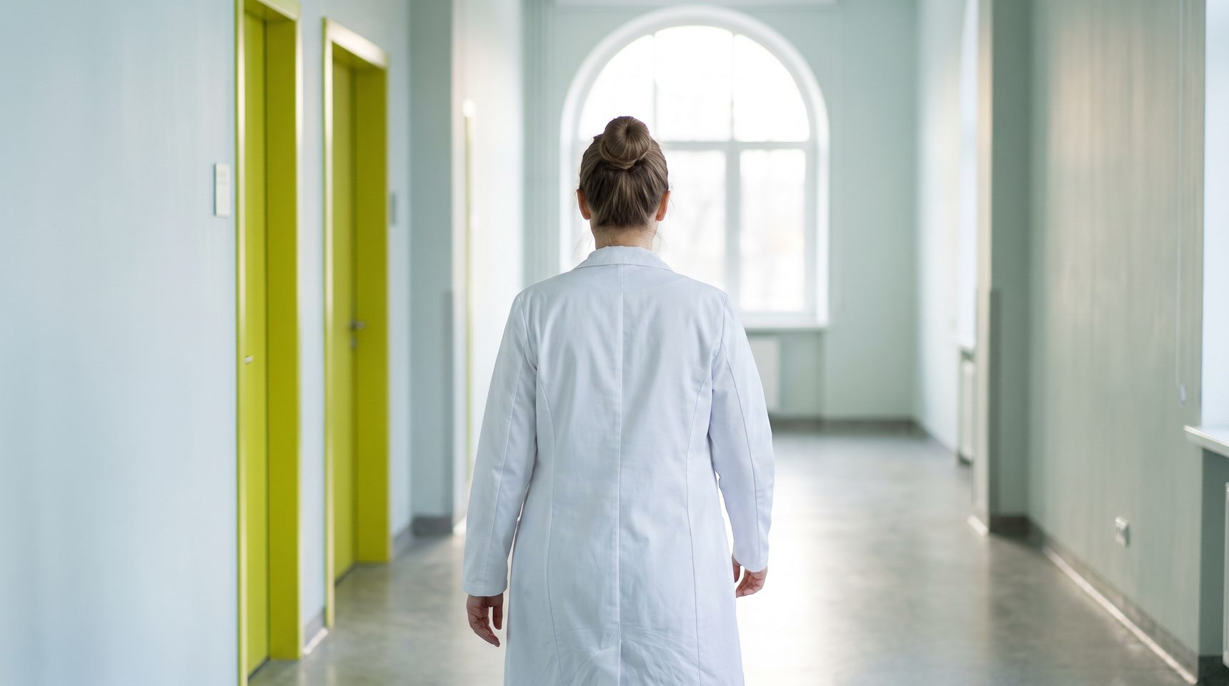 A woman in a white medical coat, seen from behind, walks toward a tall arched window at the end of a bright, airy corridor, two doors along the corridor painted yellow-green.