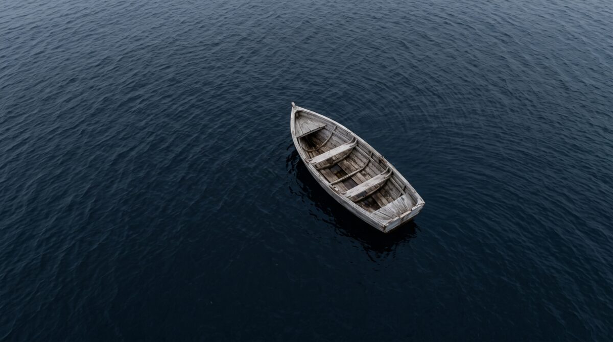 A single empty wooden rowing boat adrift on dark open water, seen from above, with nobody aboard and nothing in it.