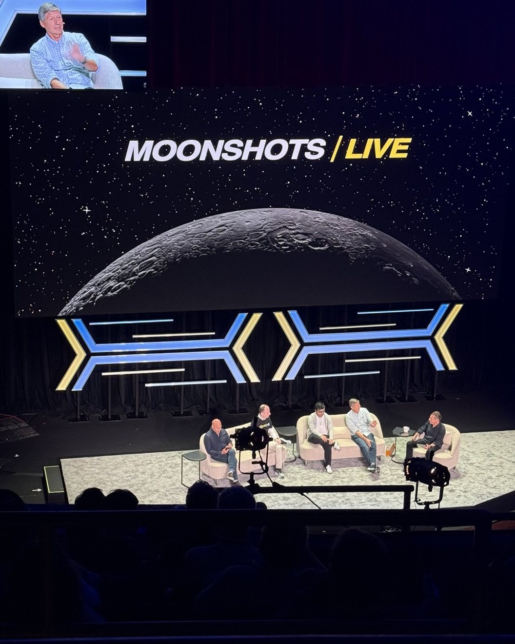 The Moonshots Live stage at the United Theater, seen from the seats: a crescent moon on the big screen, a panel of speakers on white sofas below it