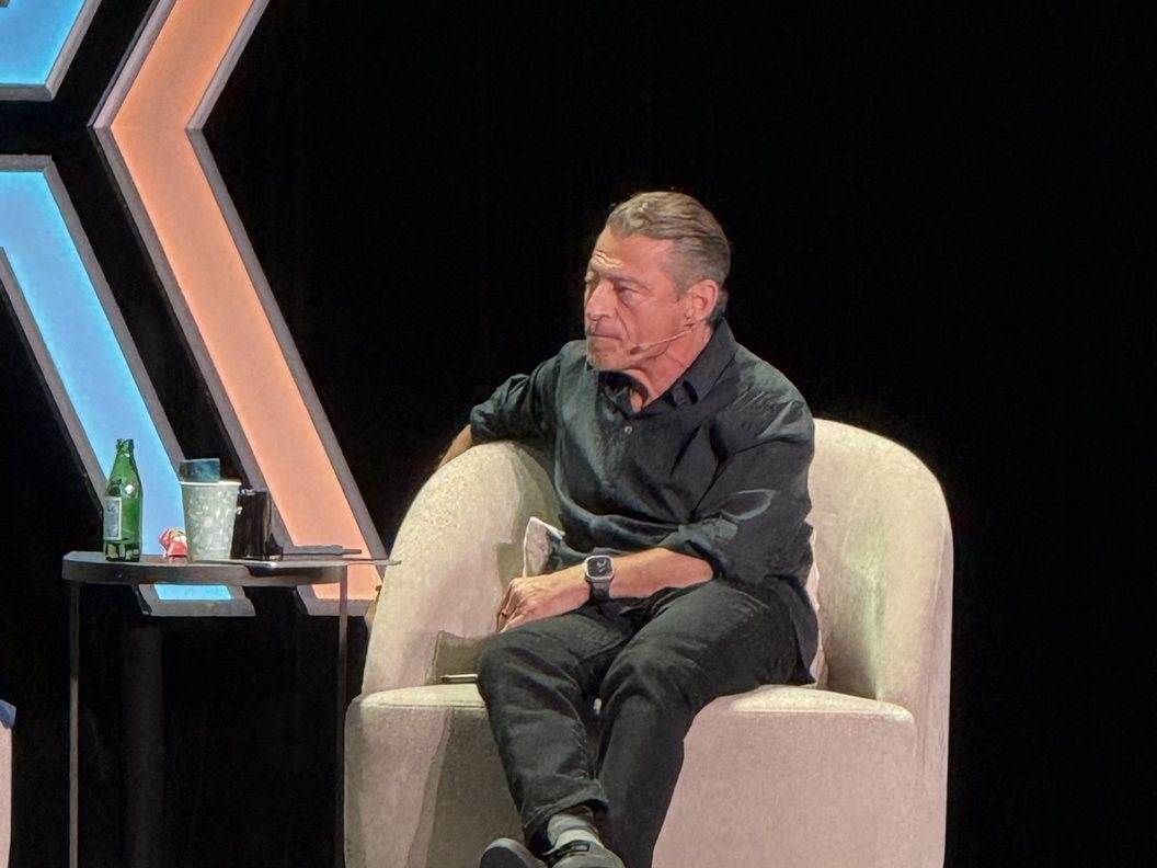 A speaker in a black shirt sits in a cream armchair on the Moonshots Live stage, a glass of water on the side table